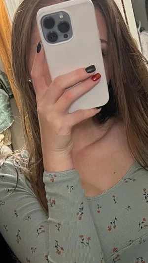 Woman with long brown hair in floral top taking mirror selfie indoors.