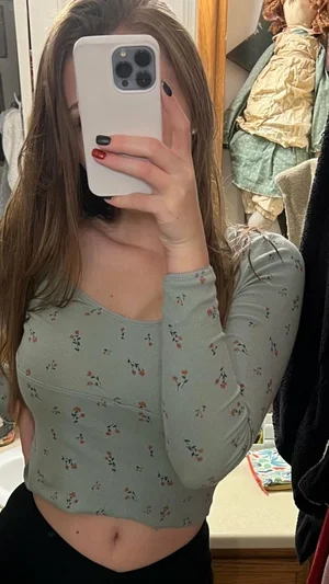 Woman in floral crop top and black bottoms taking mirror selfie in bedroom.