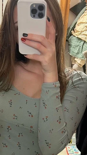 Woman with long brown hair in floral top taking mirror selfie indoors.