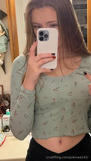 Woman in floral crop top and black pants taking mirror selfie in bathroom.