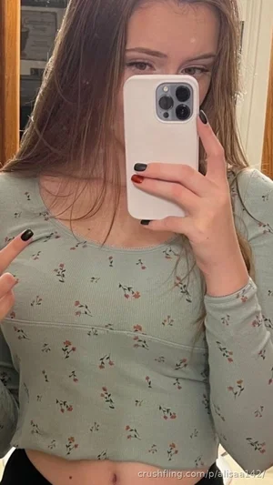 Woman with long brown hair in a floral crop top taking mirror selfie indoors.