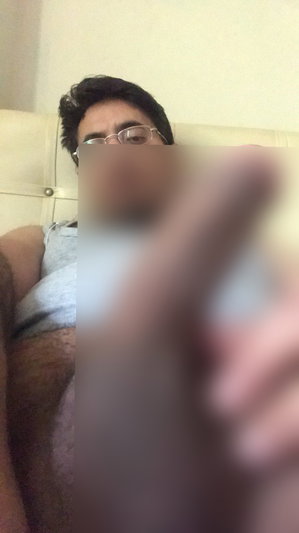 Man with glasses lying on bed, wearing gray tank top, holding phone for selfie.