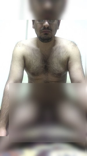 Shirtless man with glasses and beard, chest hair visible, sitting indoors.