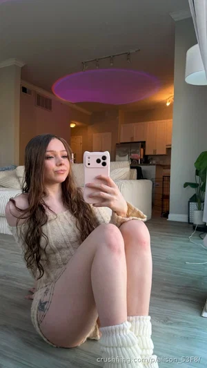 Woman with long brown hair in a light-colored dress sits on floor, taking mirror selfie in living
