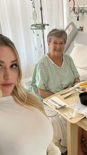 Blonde woman in white top takes selfie with older woman in hospital gown in hospital room.