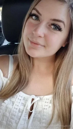 Woman with long blonde hair in white lace top, sitting in car.
