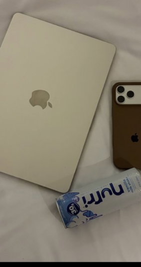 White laptop, brown phone, and Nutri-Go drink on white sheets.