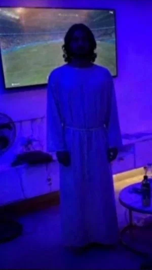Person in long white robe standing in dimly lit room with TV showing sports.