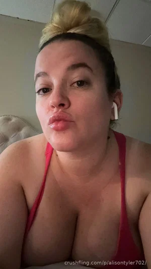 Woman with blonde hair in bun, wearing red tank top, making pouty face in bedroom selfie.