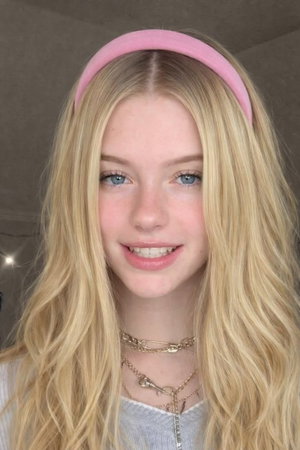 Blonde woman with blue eyes wearing pink headband and layered necklaces, indoor setting.