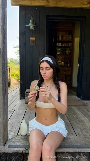 Woman in beige top and white shorts sits on wooden porch, holding small flowers.