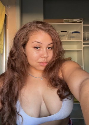 Woman with long brown hair in light blue top, taking selfie indoors.