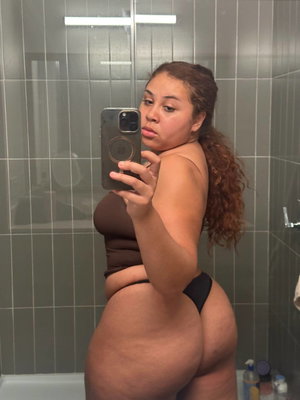 Woman in black thong and brown top taking mirror selfie in tiled bathroom.