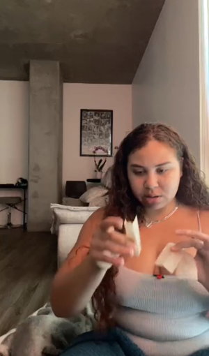 Curvy woman with curly hair in knit top and jeans holding fabric to chest in living room.