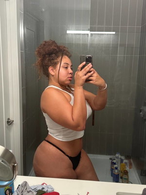 Curvy woman in white top and black thong taking bathroom mirror selfie.