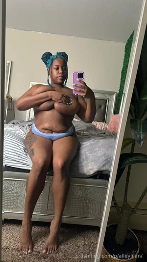 Woman with blue braided hair in light blue underwear, sitting on bed, taking mirror selfie.