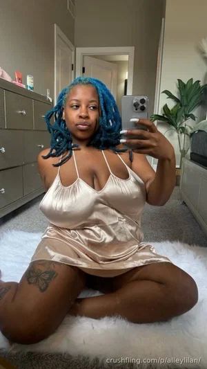 Woman with blue braids in satin slip, sitting on white rug in bedroom.
