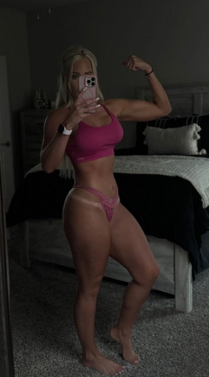 Blonde woman in pink lingerie taking mirror selfie in bedroom.