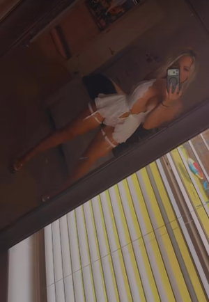 Woman with blonde hair in white outfit taking mirror selfie in bedroom.