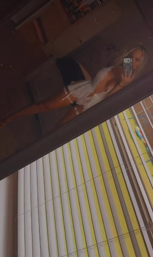 Woman with blonde hair in white top and black shorts takes mirror selfie in indoor setting.