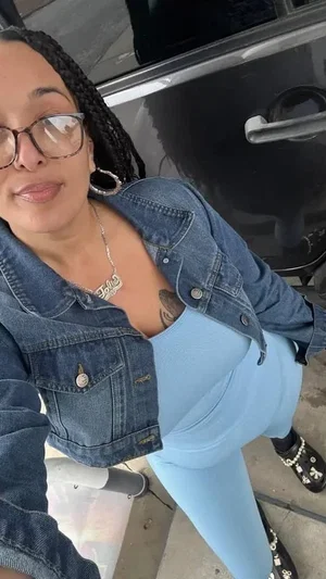 Woman with braids and glasses in denim jacket and blue outfit, posing indoors.