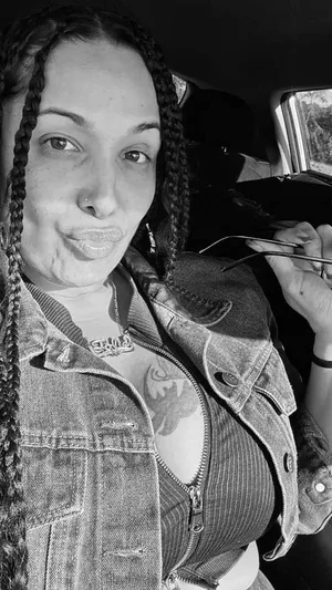 Woman with braids in denim jacket and ribbed top, sitting in car, holding glasses.