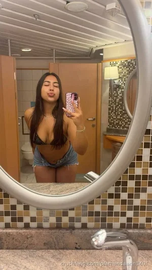 Woman in black lace bra and denim shorts taking mirror selfie in bathroom.