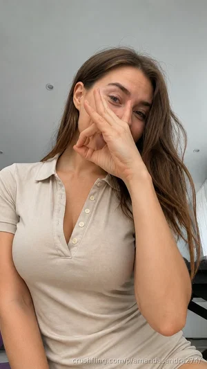 Woman with long brown hair in a beige polo shirt, covering her face partially with her hand indoors.