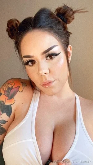 Woman with dark hair in buns, tattoo on shoulder, wearing white tank top, indoor selfie.