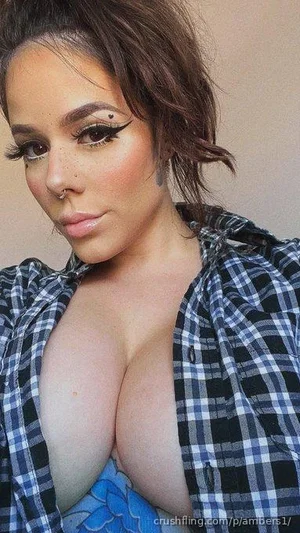 Woman with dark hair and makeup, wearing open plaid shirt over blue top, indoors.