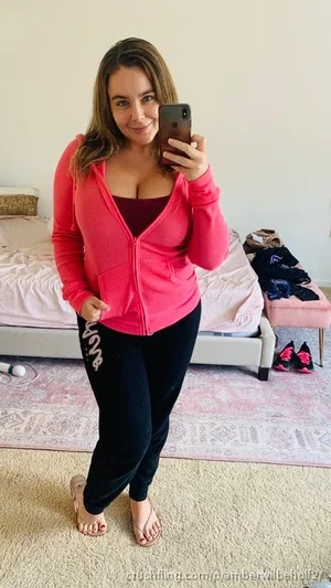 Woman in pink hoodie and black pants takes mirror selfie in bedroom.