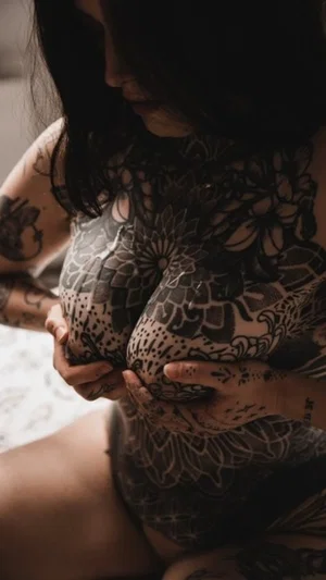 Woman with full-body tattoos holding breasts, indoor setting.