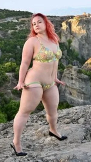 Woman with red hair in patterned bikini and black heels standing on rocky terrain outdoors.