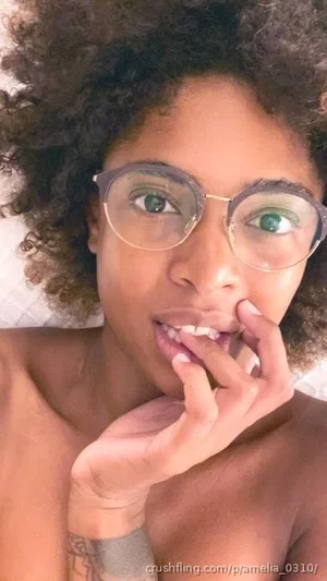 Woman with curly hair and glasses, bare-chested, hand near mouth, indoor setting.