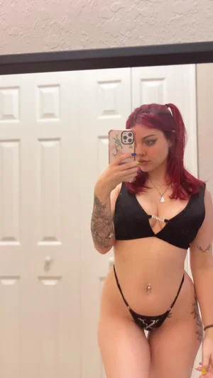 Curvy redhead in black lingerie with tattoos in bedroom mirror selfie.