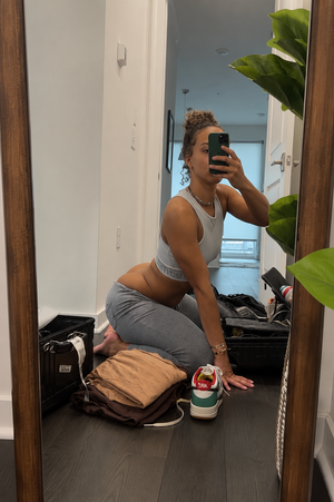 Woman in gray sports bra and leggings takes mirror selfie in hallway, packing suitcase.