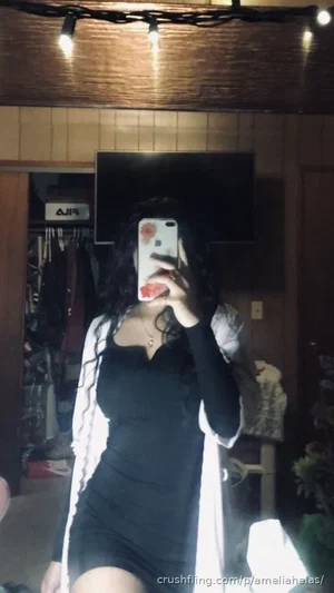 Woman in black dress and white cardigan taking mirror selfie in dimly lit room with string lights.