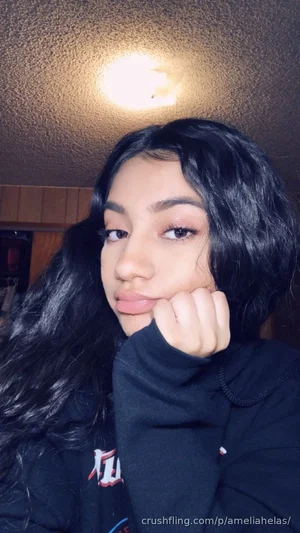 Woman with long black hair in a black hoodie, posing indoors under ceiling light.