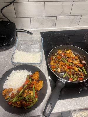 Kitchen counter with cooked meal, rice in container and on plate, stir-fry in pan.