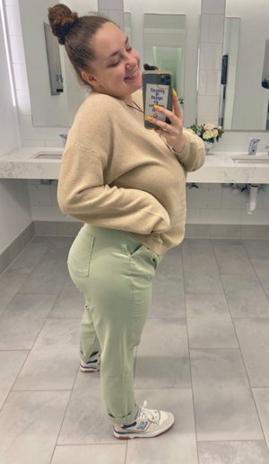 Woman in beige sweater and green pants taking mirror selfie in bathroom.