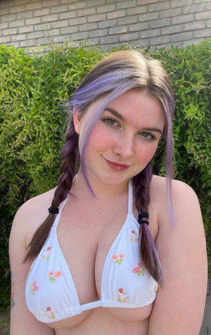 Woman with purple hair in white bikini top outdoors by bushes.