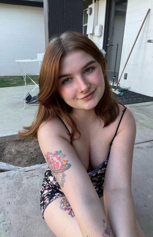 Red-haired woman with tattoos in floral top sitting outdoors.