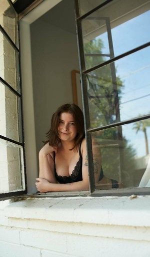Brunette woman in black lace bra leans out window.