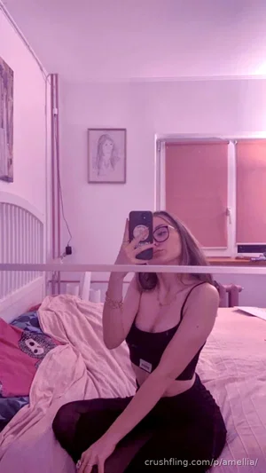 Woman in black crop top and pants sits on bed, taking mirror selfie in bedroom with pink lighting.