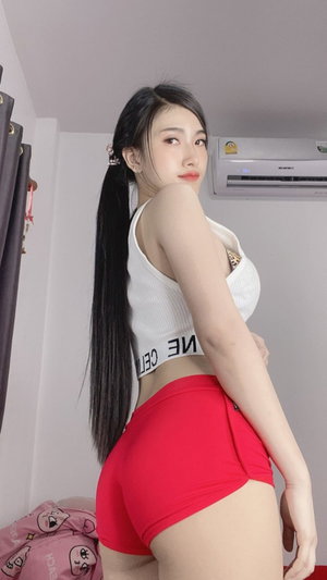 Asian woman with long ponytail in white top and red shorts indoors.