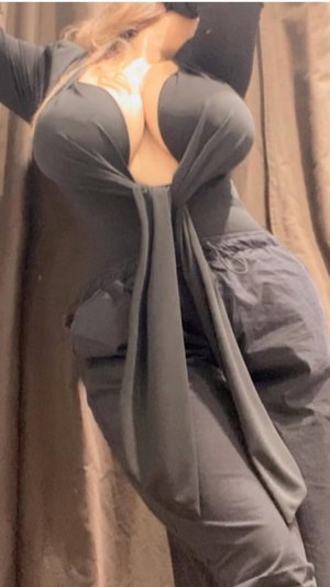 Woman in dark gray outfit with deep back cut, posing indoors near curtain.