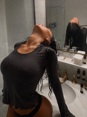 Woman with wet hair in black long-sleeve top and thong, kneeling in bathroom mirror selfie.