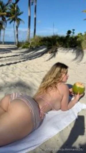 Woman in pink bikini lying on beach towel, holding coconut, palm trees in background.