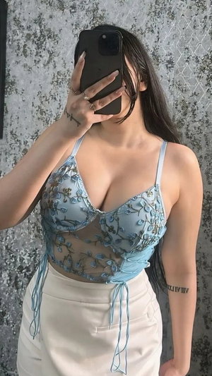 Woman in blue lace top and beige skirt, holding phone to face, standing against textured wall.