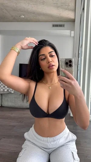 Woman in black bra and gray sweatpants taking mirror selfie indoors.